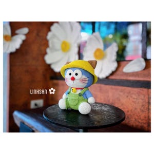 May include: A handmade blue and white crochet doll wearing a yellow hat with brown ears and green overalls. The doll has a red nose and pink whiskers. The doll is sitting on a black table. The text "LINKHSAN" is visible.
