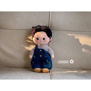 May include: A handmade crochet doll with dark hair and a light complexion. The doll wears a gray sweater and blue overalls with star accents. The doll is sitting on a gray couch. The text "LINHSAN" is visible.