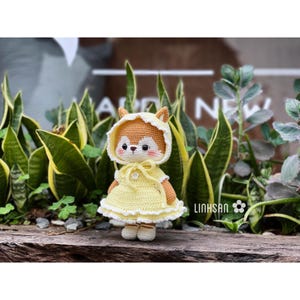Könnte beinhalten: Eine handgefertigte Häkelfuchs-Puppe mit gelbem Kleid und passender Kapuzenhaube. Der Fuchs hat ein weißes Gesicht, rosa Wangen und braune Ohren. Die Puppe steht vor grünen Pflanzen.