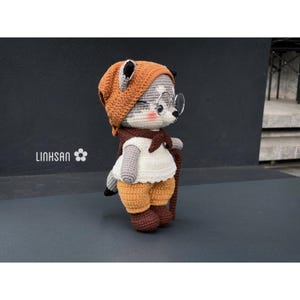 Könnte beinhalten: Gehäkelte Puppe eines Waschbären mit braunem Hut, Brille und Schal. Der Waschbär trägt ein weißes Hemd, eine braune Hose und hält einen Wanderstock. Der Waschbär hat ein grau-weißes Gesicht mit rosa Wangen. Das Wort "LINHSAN" ist sichtbar.