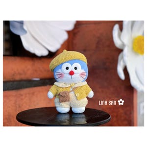 May include: A handmade crochet doll of a blue cat-like character wearing a yellow beret, jacket, and shorts. The doll has red eyes, pink cheeks, and a small brown bag. The doll is standing on a dark brown surface.