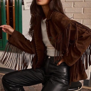 May include: A brown suede jacket with long fringe detailing on the sleeves. The jacket is paired with black leather trousers. The model is wearing black sunglasses. The brand logo "COZY CLASSY CHIC" is visible in the top left corner.