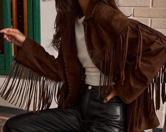 Könnte beinhalten: Braune Wildlederjacke mit langen Fransen an den Ärmeln. Die Jacke wird mit einer schwarzen Lederhose kombiniert. Das Model trägt eine schwarze Sonnenbrille. Das Markenlogo "COZY CLASSY CHIC" ist oben links sichtbar.