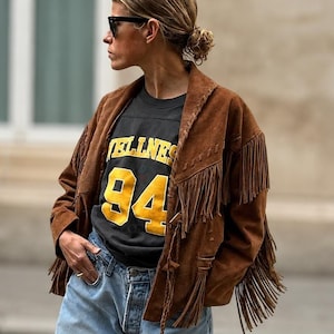 Puede incluir: Una chaqueta de ante marrón con largos flecos en el pecho y las mangas, sobre una camiseta negra con el texto amarillo "WELLNESS 94" y vaqueros azul claro. La chaqueta tiene un cuello estructurado y mangas largas.