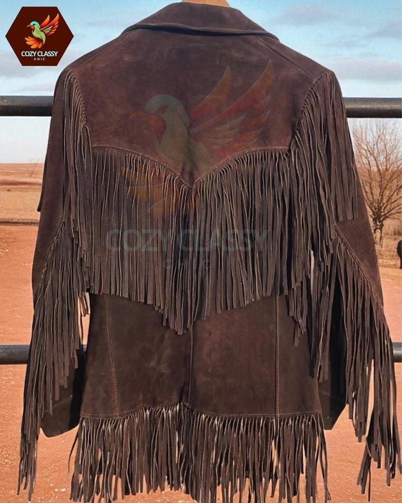 K&ouml;nnte beinhalten: Braune Wildlederjacke mit langen Fransen an den &Auml;rmeln und am R&uuml;cken. Auf der R&uuml;ckseite der Jacke befindet sich ein gemaltes Adlerdesign. Die Jacke hat einen Kragen und einen Knopfverschluss. Das Cozy Classy Chic-Logo ist oben links sichtbar.