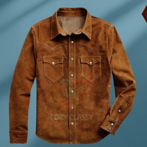 Pode incluir: Uma camisa de camurça marrom com mangas compridas e fechamento com botões. A camisa tem dois bolsos no peito com fechamento com botões e uma gola pontiaguda.