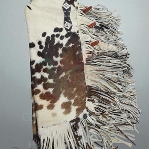 Puede incluir: Chaps de piel de vaca con un canesú de cuero negro y adornos plateados. Los chaps presentan un estampado de manchas marrones y blancas y largas flecos. Se incluyen correas de cuero marrón. Estos chaps están diseñados para montar a caballo.