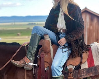 May include: A person wearing a cowboy hat, a brown suede jacket with fringe, blue jeans, and green cowboy boots, sitting on a horse. The saddle is brown leather, and the background shows a rural landscape.