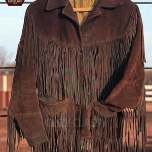 K&ouml;nnte beinhalten: Braune Wildlederjacke mit langen Fransen an der Vorderseite, den &Auml;rmeln und den Taschen. Die Jacke hat einen Kragen und zwei Vordertaschen. Die Jacke h&auml;ngt an einem Holzb&uuml;gel.