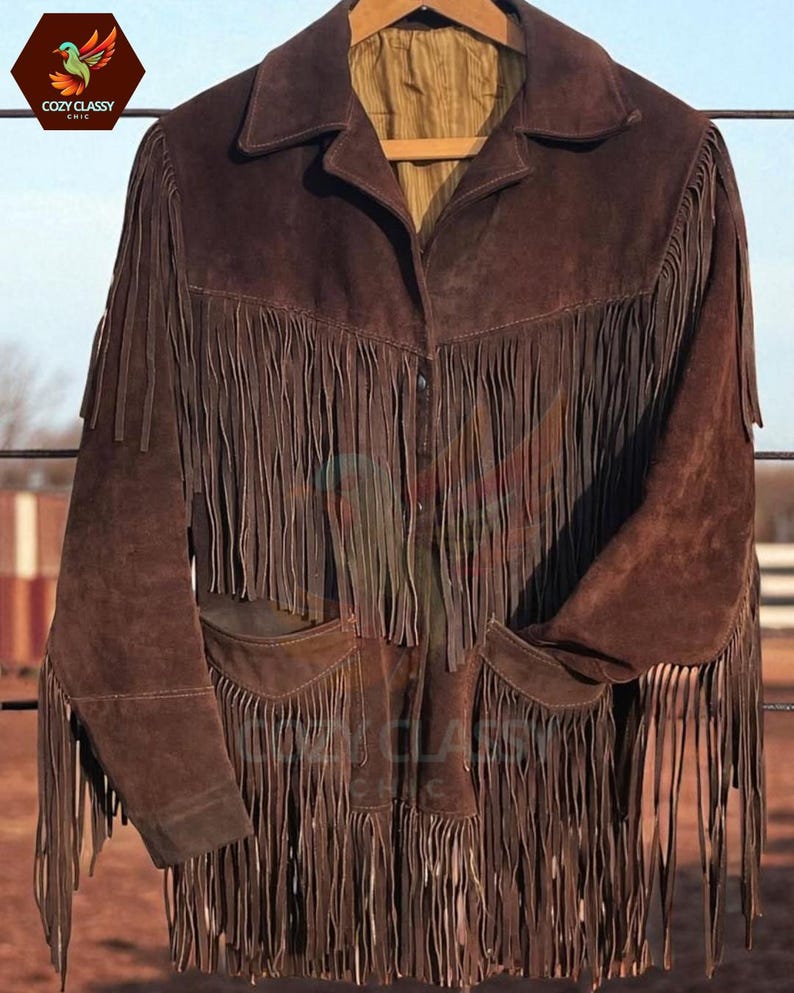 K&ouml;nnte beinhalten: Braune Wildlederjacke mit langen Fransen an der Vorderseite, den &Auml;rmeln und den Taschen. Die Jacke hat einen Kragen und zwei Vordertaschen. Die Jacke h&auml;ngt an einem Holzb&uuml;gel.