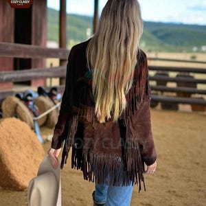 K&ouml;nnte beinhalten: Braune Wildlederjacke mit Fransen. Die Jacke wird mit blauen Jeans und Cowboy-Stiefeln getragen. Ein cremefarbener Cowboyhut wird in der Hand gehalten. Der Hintergrund umfasst einen Holzzaun und eine l&auml;ndliche Landschaft.