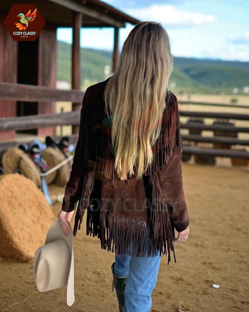 K&ouml;nnte beinhalten: Braune Wildlederjacke mit Fransen. Die Jacke wird mit blauen Jeans und Cowboy-Stiefeln getragen. Ein cremefarbener Cowboyhut wird in der Hand gehalten. Der Hintergrund umfasst einen Holzzaun und eine l&auml;ndliche Landschaft.