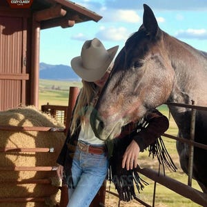 K&ouml;nnte beinhalten: Eine Person mit hellem Cowboyhut, schwarzer Jacke mit Fransen und blauer Jeans, die sich an einen Metallzaun neben einem braunen Pferd lehnt. Die Szene spielt sich im Freien mit einer Scheune und einem Feld im Hintergrund ab.