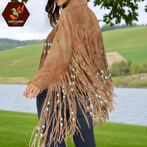 Puede incluir: Chaqueta de ante marrón con largos flecos y detalles de cuentas. La chaqueta tiene cuello y se lleva sobre pantalones oscuros. El logotipo "Cozy Classy Chic" es visible en la esquina superior izquierda.