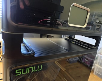 May include: A black 3D printer with the brand name SUNLU in green lettering. The printer has a sleek, modern design with a rectangular screen and a ridged platform. The printer is on a wooden surface.