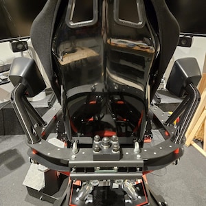 Op de afbeelding: Zwarte race-simulatorstoel met een zwart metalen frame en rode accenten. De stoel heeft een hoge rugleuning met twee rechthoekige uitsparingen. Het frame heeft een reeks bouten en moeren.