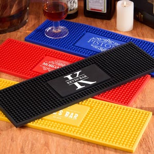 以下が含まれることがあります： 黒、赤、青、黄色の長方形のバーマットのコレクション。各マットは、テクスチャ加工された表面と、バー名が記載された長方形のラベルが付いています。こぼれたものをキャッチし、表面を保護するように設計されています。