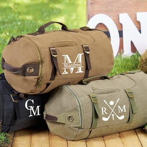 May include: Three canvas duffel bags with personalized embroidery. The first bag is brown with the name "Manfred" and a large "M". The second bag is black with the initials "GM". The third bag is green with the initials "RM" and a golf club and ball graphic.