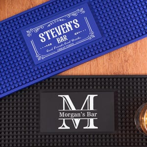 Puede incluir: Dos alfombrillas rectangulares para bar. La alfombrilla superior es azul con el texto "STEVEN'S BAR" en blanco. La alfombrilla inferior es negra con el texto "Morgan's Bar" en blanco. Un vaso con líquido dorado está sobre la alfombrilla negra.