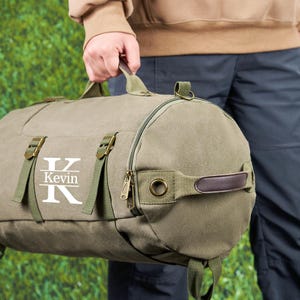 May include: A green canvas duffel bag with a brown leather handle and a zipper closure. The bag has a white monogram with the letter "K" and the name "Kevin" printed on it.