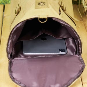 May include: A beige canvas backpack with a zippered opening, revealing a purple lining and a black tablet computer inside.