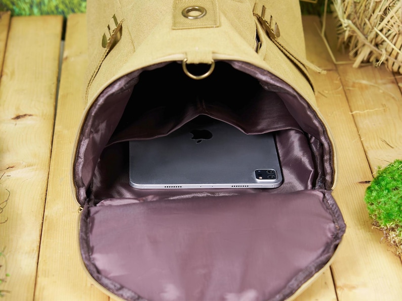 May include: A beige canvas backpack with a zippered opening, revealing a purple lining and a black tablet computer inside.