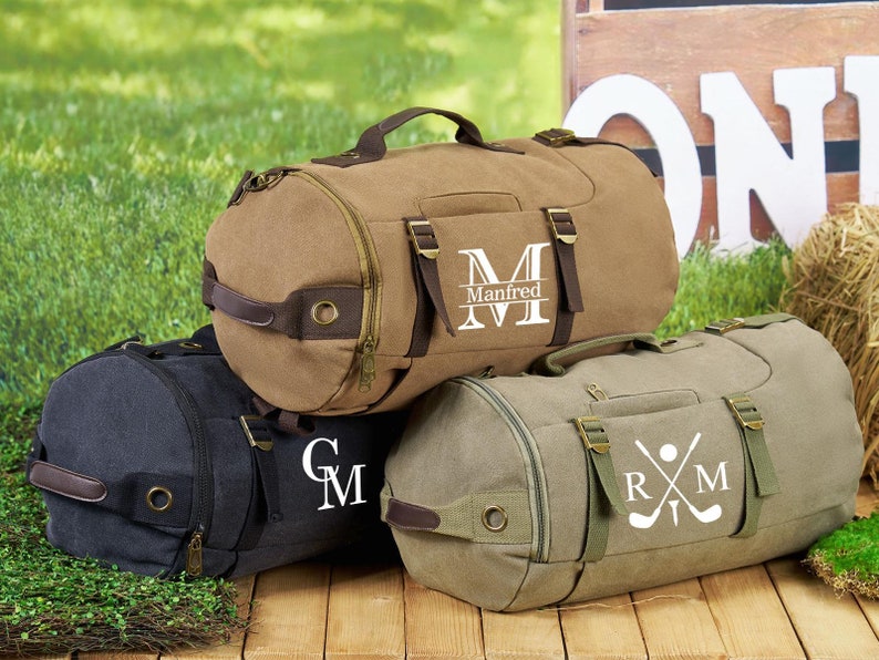May include: Three canvas duffel bags with personalized embroidery. The first bag is brown with the name "Manfred" and a large "M". The second bag is black with the initials "GM". The third bag is green with the initials "RM" and a golf club and ball graphic.