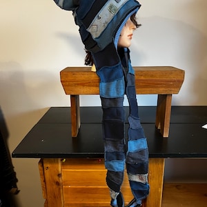 Peut inclure: Ensemble chapeau et écharpe en patchwork dans les tons de bleu, noir et gris. Le chapeau a un sommet pointu et est fait de divers carrés de tissu. L'écharpe est longue et pend, également faite de patchwork. L'ensemble est présenté sur une tête de mannequin.