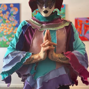 Op de afbeelding: Een patchwork trui in tinten van turquoise, paars, roze en lavendel. De trui heeft een hoge kraag met ruches en uitlopende mouwen. Een persoon draagt een masker en een hoge hoed.