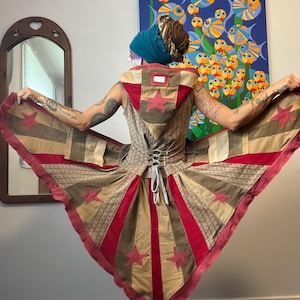 May include: A unique, handmade garment featuring a patchwork design with red stars and stripes. The bodice has a corset-style back, and the skirt flares out. The color palette includes tan, red, and brown tones. The garment is a distinctive piece of wearable art.