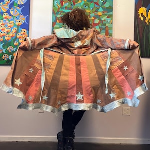 May include: A long, flared coat featuring a sunburst pattern of alternating brown, tan, and coral-pink vertical panels. The hem is trimmed with a metallic silver border and decorated with multiple light blue star-shaped appliqués throughout.