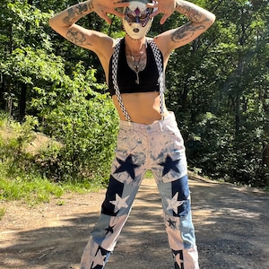 May include: A person wearing a black crop top, white pants with star patches, and black suspenders with a checkered pattern. The person is also wearing a mask and a head wrap. The background is a dirt path and trees.