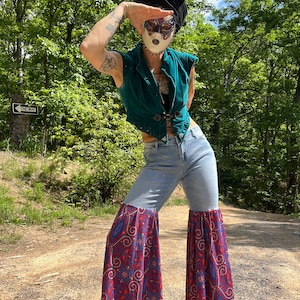 May include: A person wearing a teal velvet waistcoat, light blue jeans with a red, blue and white patterned fabric sewn onto the bottom of the legs, and a black headscarf. The person is wearing a white mask with purple and gold accents. The person is standing on a dirt road with trees in the background. A sign that reads "One Way" is visible in the background.