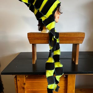 Puede incluir: Un sombrero caprichoso, hecho a mano, con una larga cola colgante. El sombrero presenta rayas negras y amarillo neón alternadas, creando un diseño audaz y llamativo. La tela parece ser un mosaico de cuadrados, lo que se suma a su atractivo único y artesanal. El sombrero se exhibe en una cabeza de maniquí.