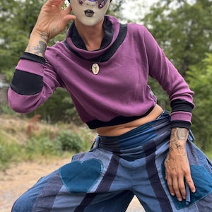 May include: A person wearing a purple turtleneck sweater with black trim, blue and white striped pants with heart-shaped patches, and a white mask with a butterfly design.