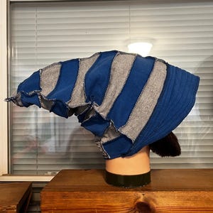 May include: A slouchy beanie hat constructed from alternating vertical panels of blue and heather gray fabric. The design features exposed seams with a raw-edge aesthetic, creating a textured appearance. The hat has a wide, ribbed blue brim.