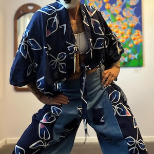 Op de afbeelding: Een marineblauwe kimono-achtige jas en bijpassende broek met een wit bladerpatroon en roze accenten. De broek heeft denim patches. De persoon draagt een witte top en een decoratief masker.