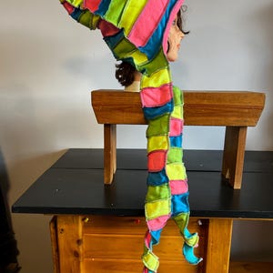 Pode incluir: Um chapéu de patchwork colorido com uma longa echarpe, exibido em uma cabeça de manequim. O chapéu e a echarpe apresentam listras vibrantes de rosa, verde, amarelo e azul, costuradas com costuras visíveis. O chapéu tem uma ponta.
