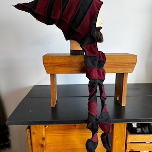 May include: A handmade, striped hat with a long, spiralling tail. The hat is black and burgundy, with a pointed tip and a hood. The hat is displayed on a mannequin head. The hat is made of a soft, textured fabric.