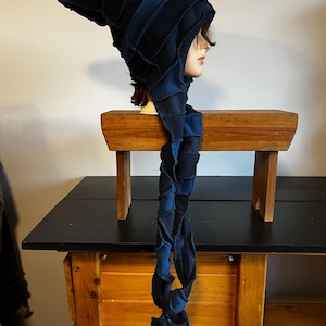 Peut inclure: Un chapeau bleu foncé de style patchwork avec une longue écharpe tressée. Le chapeau a un sommet pointu et est présenté sur une tête de mannequin. L'écharpe pend, mettant en valeur le design et la texture uniques du chapeau. Le chapeau est fait d'une matière douce.