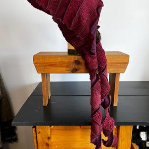May include: A burgundy-colored, handmade hat with a long, spiraling scarf. The hat features a pointed top and is constructed from a combination of textured and ribbed fabrics. The scarf has a unique, twisted design with decorative elements.