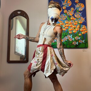 Op de afbeelding: Een persoon draagt een witte tanktop en een patchwork rok met rode en bloemenpatronen. De persoon draagt ook een masker en heeft tatoeages op zijn armen. Een schilderij van kleurrijke vissen hangt aan de muur achter hen.