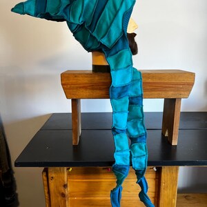 May include: A handmade, patchwork hat and scarf set in shades of teal and blue. The hat has a long, pointed tip and the scarf hangs down with two curled ends. The set is displayed on a mannequin head, showcasing the unique design and craftsmanship.