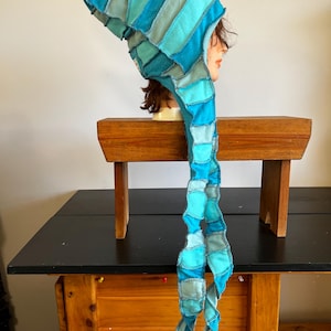 Pode incluir: Um chapéu de patchwork em tons de azul e turquesa, com uma longa cauda fluida. O chapéu é feito de pedaços de tecido retangulares costurados, criando um design texturizado. O chapéu é exibido em uma cabeça de manequim.