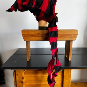 Pode incluir: Um chapéu listrado vermelho e preto com uma longa cauda torcida. O chapéu é feito de tecido e tem um topo pontudo. O chapéu é exibido em uma cabeça de manequim.