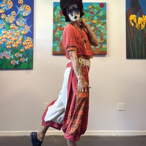 May include: A dark brown corduroy hat, a short-sleeved orange top, and loose-fitting trousers featuring a combination of solid red, white, and patterned orange fabric panels with a tie waist, paired with black slip-on shoes and a face mask.