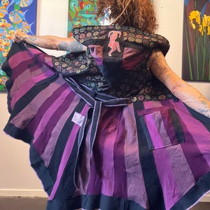May include: A hooded vest featuring a black and purple vertical striped lower section and a dark patterned upper section with a small pink figure graphic on the hood. The garment includes a tie waist, a square pocket, and a white label on the interior.