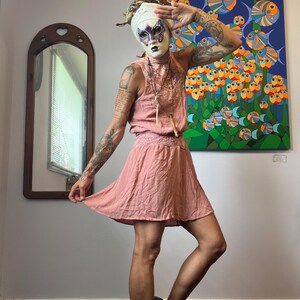 May include: A person in a pink dress with a high neckline and skirt, wearing a white head covering and a decorative mask. A necklace with a pendant is visible. A colourful painting of fish is in the background. The person is wearing black shoes.
