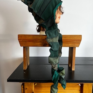 May include: A patchwork green hood with a long, spiraling scarf. The hood and scarf are made of various shades of green fabric, creating a textured and layered appearance. The hood is displayed on a mannequin head.