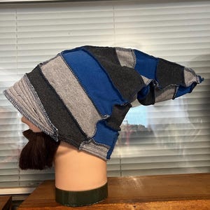 May include: A slouchy beanie hat constructed from alternating vertical panels of dark gray, light gray, and royal blue fabric. The hat features visible exposed seams with blue stitching, creating a patchwork design that tapers toward the back.